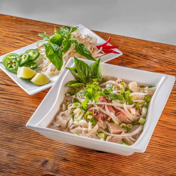 A bowl of vPho's Vietnamese pho with a side plate of fresh basil, bean sprouts, jalapeños, and lime.
