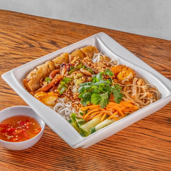 A square white bowl of vPho vermicelli noodles with spring rolls, shrimp, and vegetables.