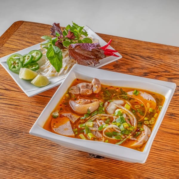A bowl of vPho's Vietnamese bun bo hue soup with a side plate of fresh herbs and vegetables.