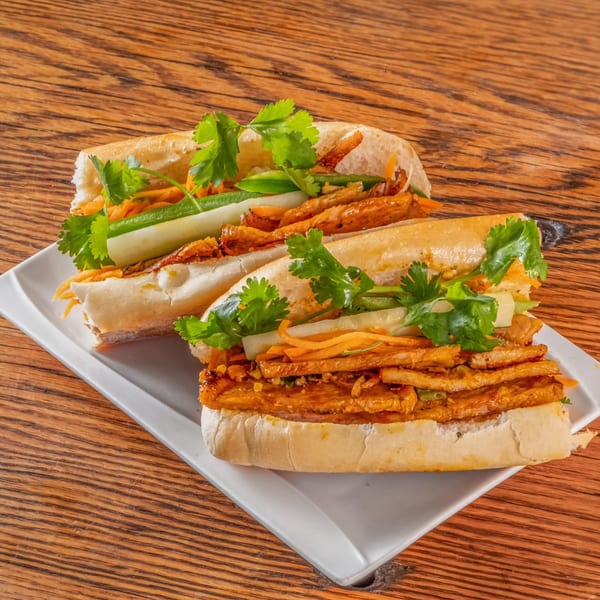 Two vPho banh mi sandwiches with grilled pork, pickled vegetables, and cilantro on a white plate.