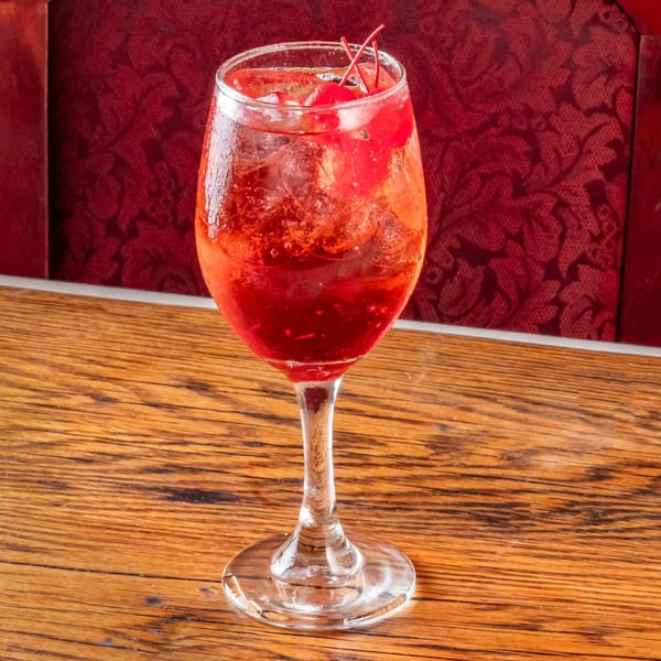 A red cocktail with ice and cherries in a wine glass on a wooden table.